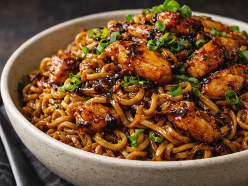 A Close Up Bowl Of Sticky Garlic Chicken Noodles Garnished With Green